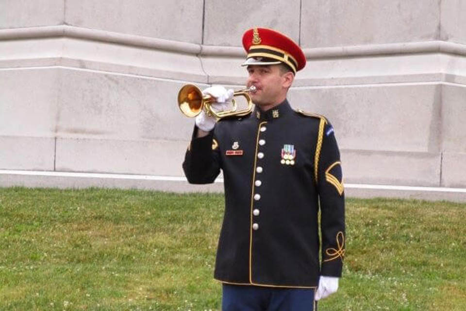 A uniformed military bugler plays a trumpet during a ceremony, wearing a red and gold cap with medals on his chest.