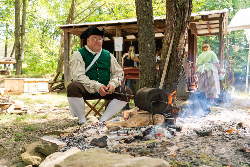 An individual in period clothing is seated while tending a campfire in an outdoor historical reenactment setting, with a wooden structure and other people in the background.