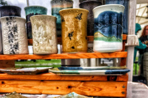 Handcrafted ceramic bowls and plates with textured blue and green glazes are displayed on a wooden shelf at a market stall.
