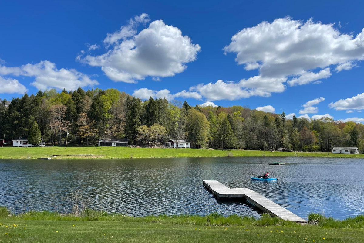 A person in a small boat on a lake under a blue sky with scattered clouds. Trees and houses are visible on the shore in the background.