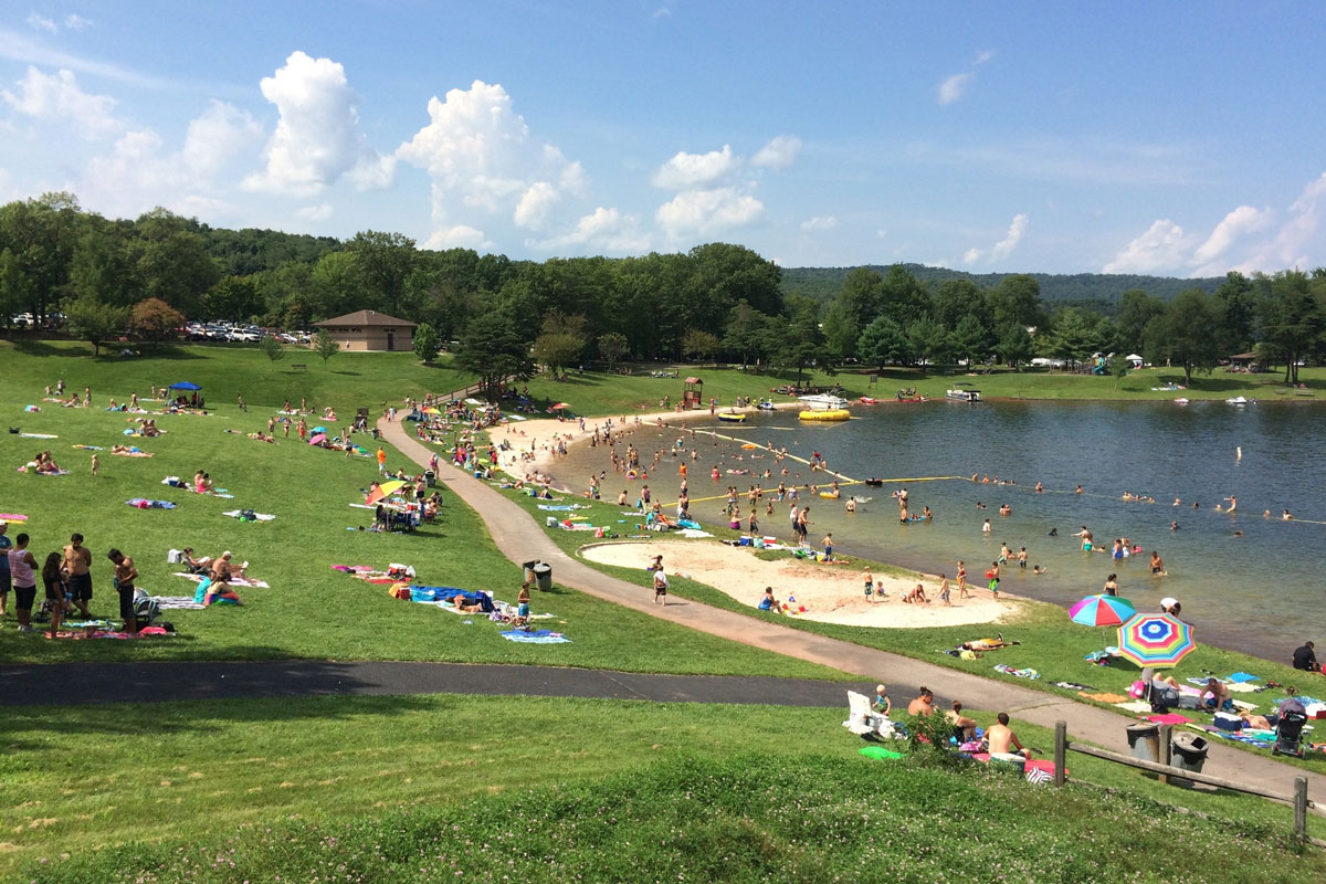 People enjoy a sunny day by a lakeside beach. Many are sunbathing on the grass, swimming, or gathered near the water. Trees and a building are visible in the background.