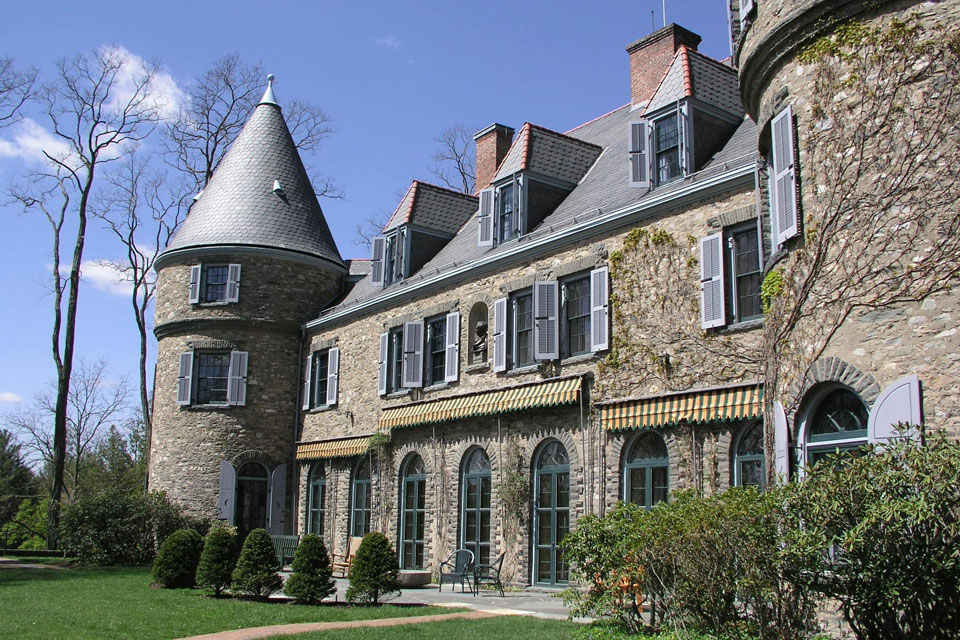 Stone mansion with a turret, multiple chimneys, and arched windows, surrounded by shrubs, under a blue sky.