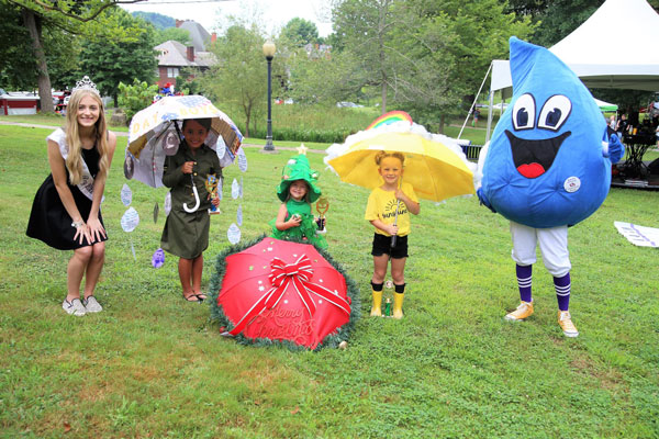 three kids holding umbrella between a water drop clown on right and a beauty pageant queen standing on left 