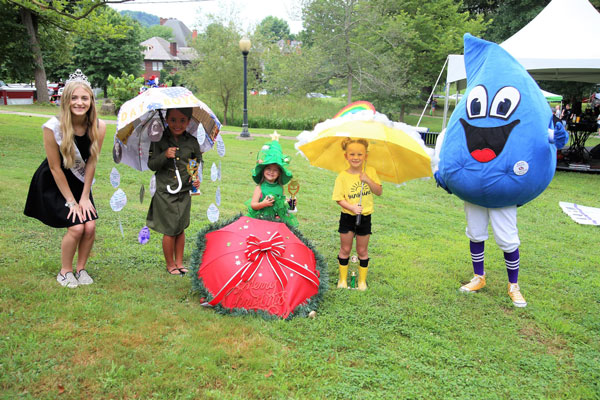 three kids holding umbrella between a water drop clown on right and a beauty pageant queen standing on left