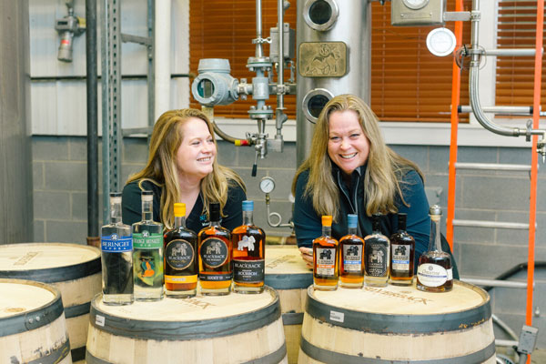 women distillers standing behind bottles place on top of a barrel