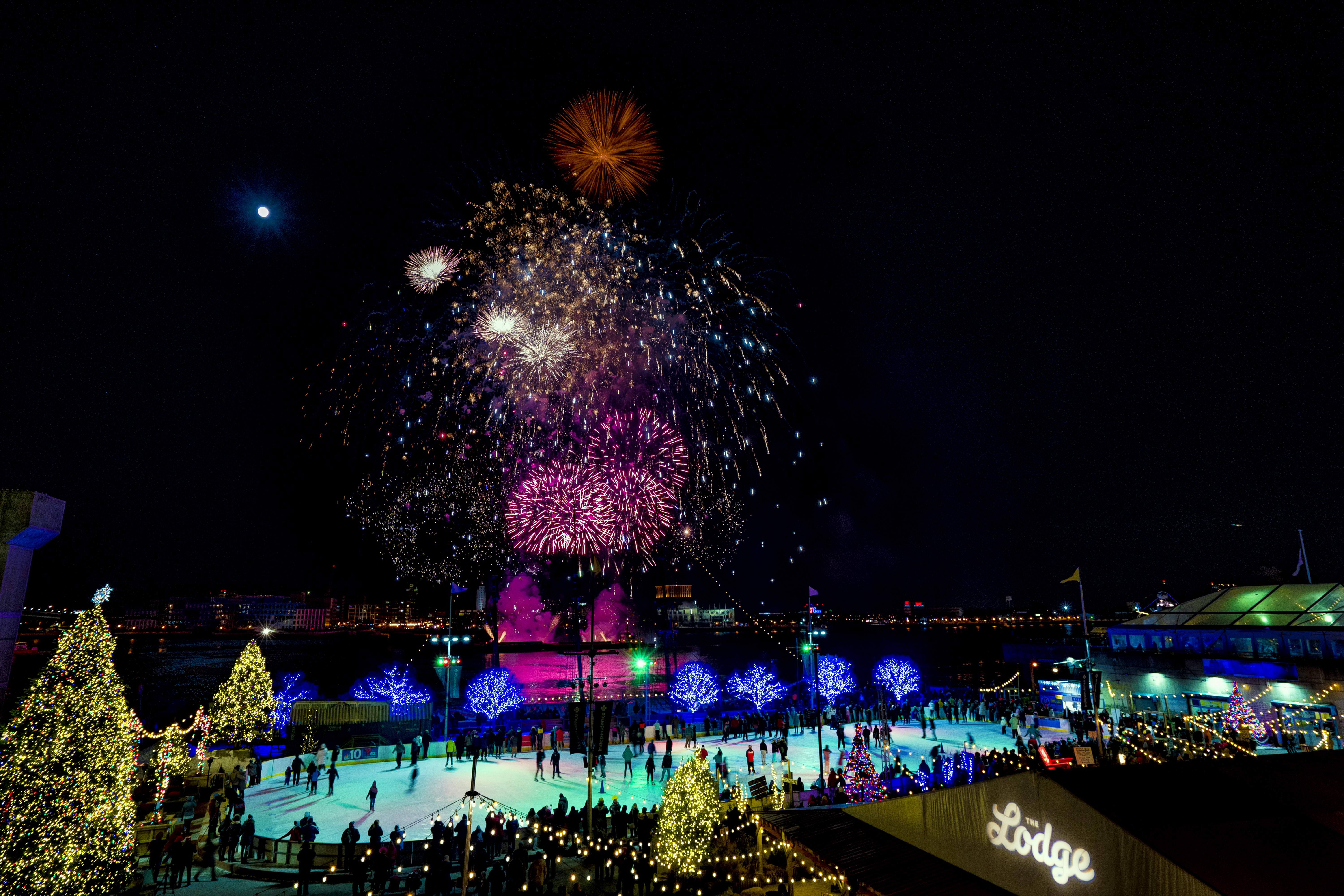 Fireworks bursting in the dark sky above people ice skating on a rink that is surrounding by lit up Christmas trees