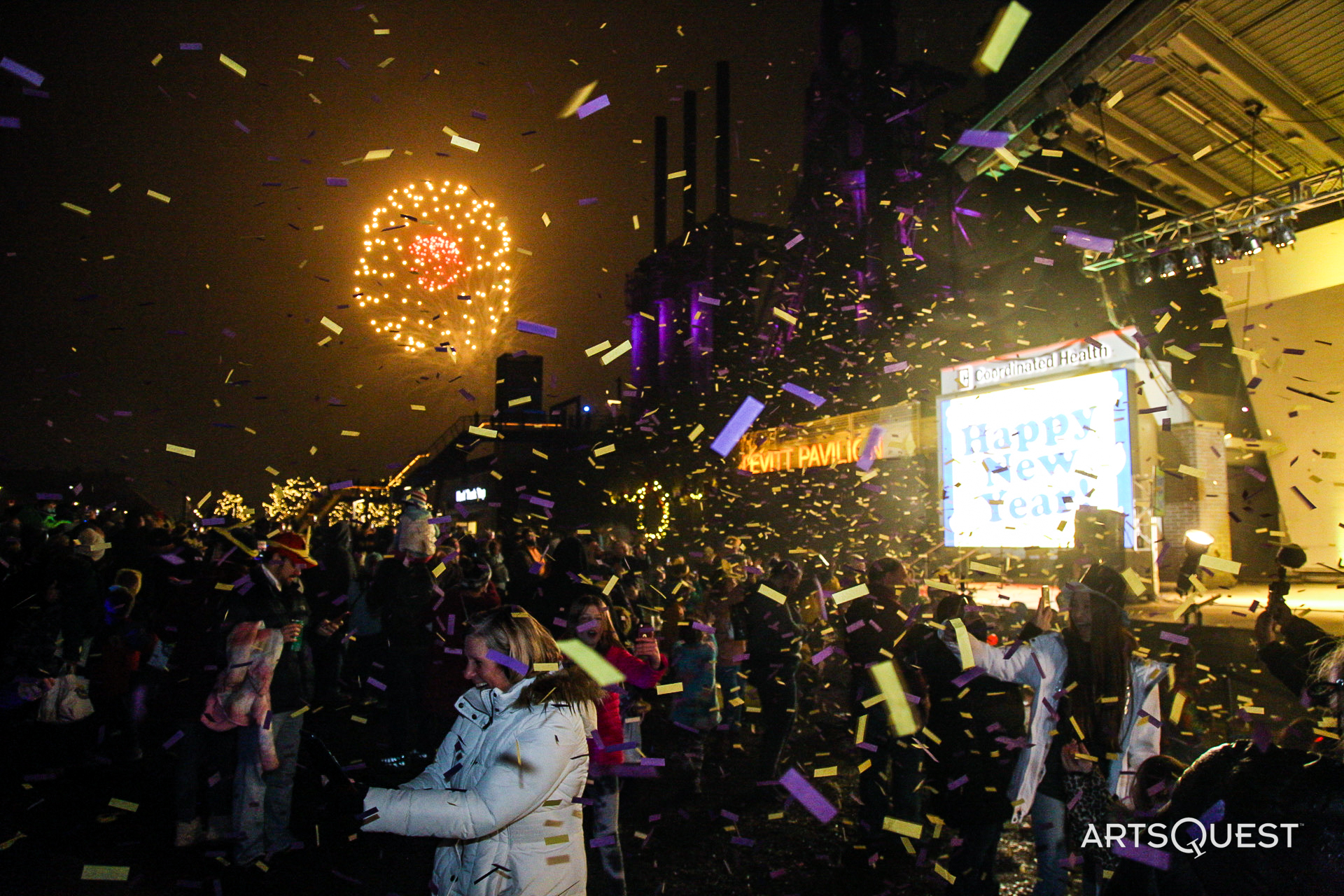People cheering with purple and yellow confetti in front of a stage with fireworks going off