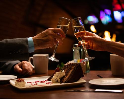  Two people toasting with champagne over dessert in a dimly lit restaurant.