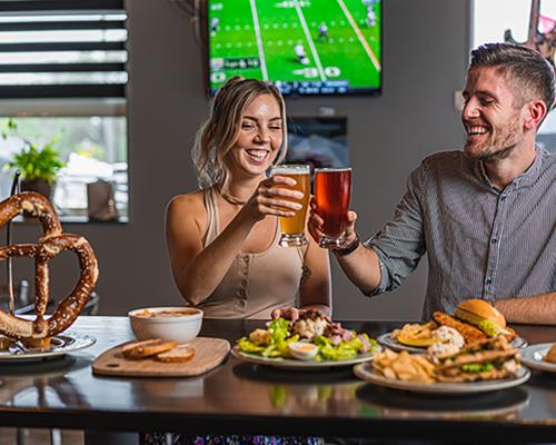 Man and woman seated with food and cheersing beers