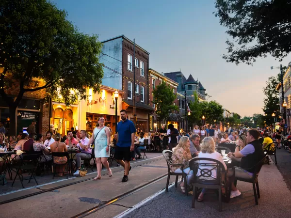 People dining at outdoor tables on a bustling street at dusk, with historic buildings and street lights illuminated