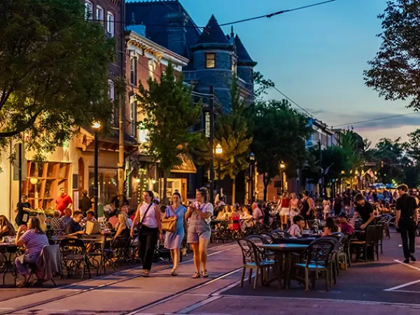 People dining at outdoor tables on a bustling street at dusk, with historic buildings and street lights illuminated